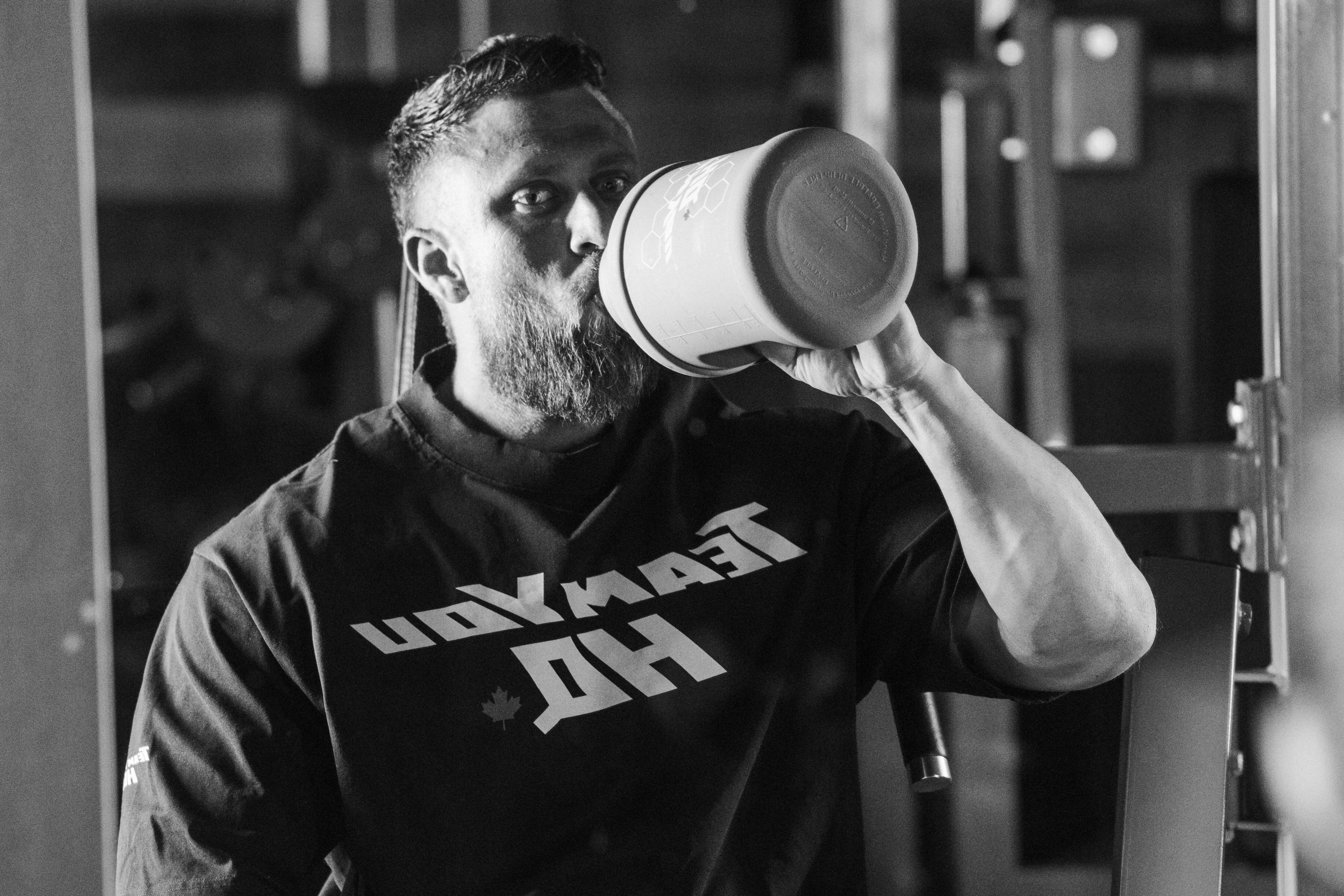 Person in a gym wearing a black t-shirt with visible branding, sitting on a bench and drinking from a large container.